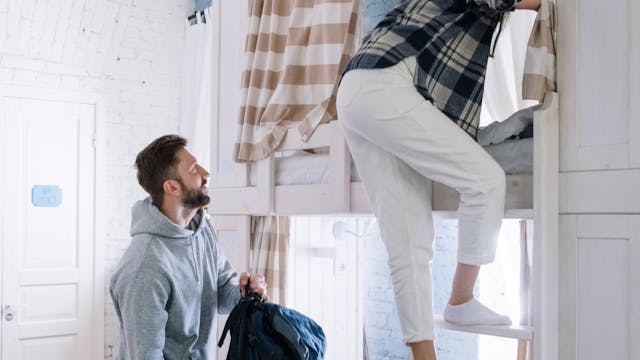 Couple packing bags in a bright, cozy hostel dormitory room with bunk beds and wooden floor.